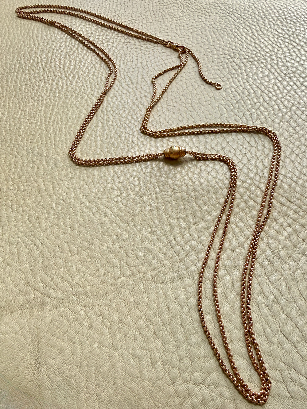 An 18k rose gold victorian chain necklace with a slider central bead detail, laid out on a textured beige leather surface.
