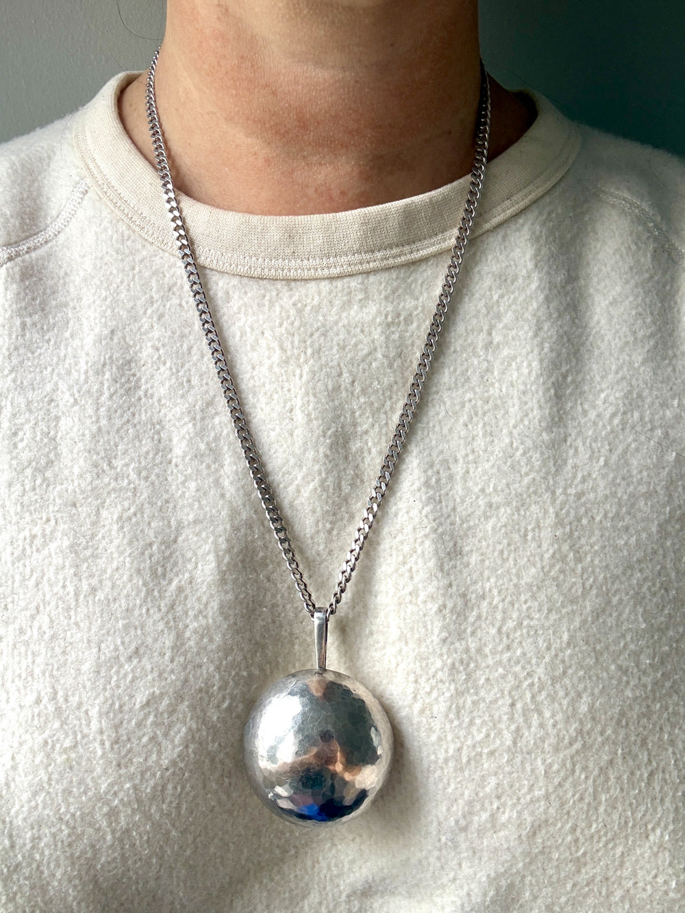 A close-up of a person wearing a silver chain necklace with a large, polished silver sphere pendant, styled with a cream-colored crewneck sweatshirt.