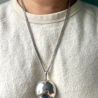 A close-up of a person wearing a silver chain necklace with a large, polished silver sphere pendant, styled with a cream-colored crewneck sweatshirt.