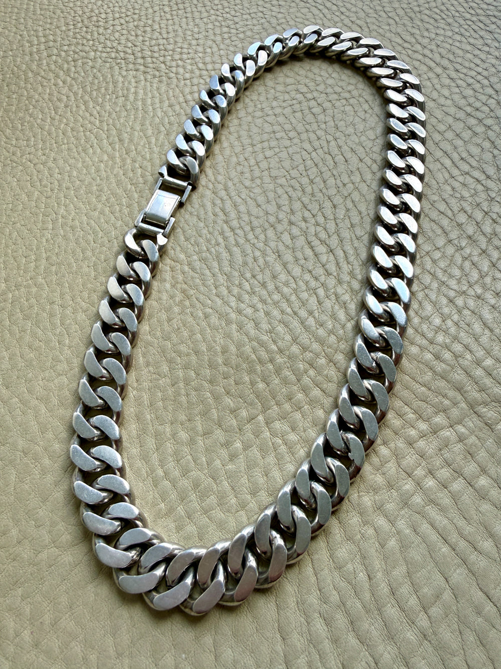 A vintage silver weighty curb link chain necklace displayed on a textured beige leather surface.