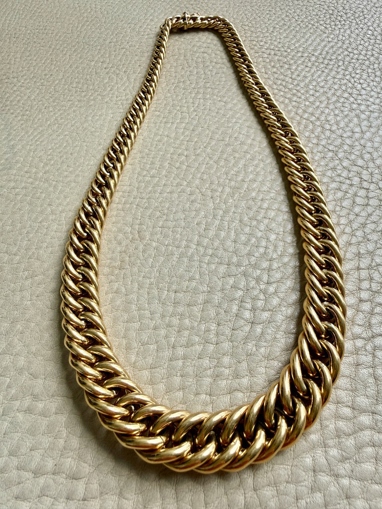 A thick, chunky 18k gold curb link chain necklace displayed on a textured off-white leather surface.