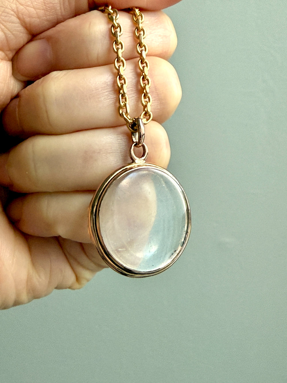 Gold necklace with a round pendant held by a hand against a light green background