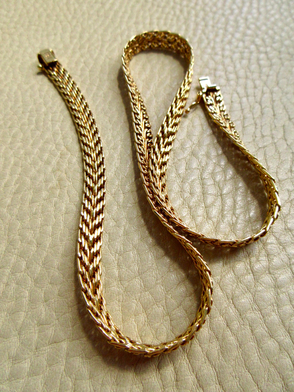 An 18k gold braided chain necklace with a box clasp, displayed on a textured beige leather surface.