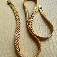 An 18k gold braided chain necklace with a box clasp, displayed on a textured beige leather surface.