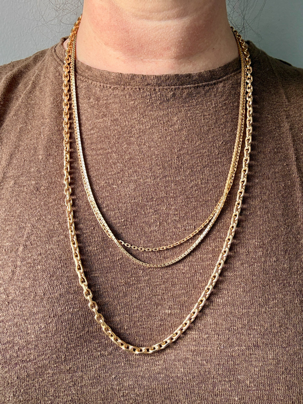 Gold chain necklace on a person wearing a brown shirt against a gray background