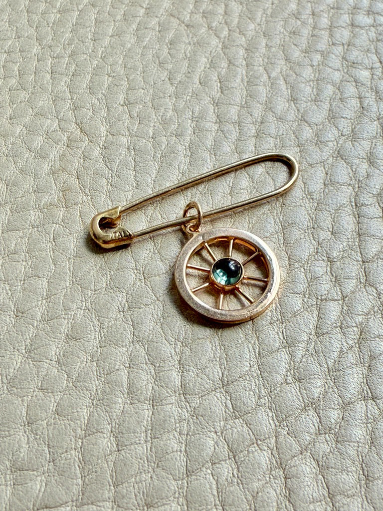 Gold safety pin with a wheel charm and blue gemstone on a textured beige background