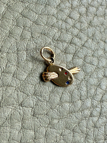 Gold charm resembling a paint palette with red, yellow, and blue dots on a textured gray background