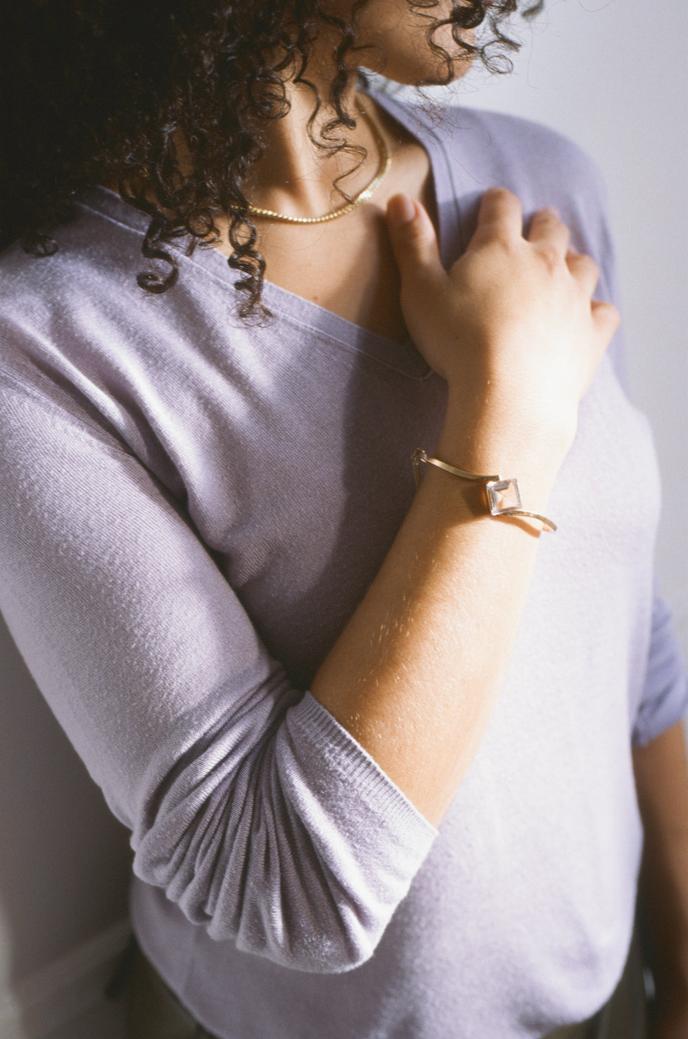 A close-up lifestyle shot of a person wearing a lavender long-sleeve knit top, a gold chain necklace, and a gold bracelet with a square bezel-set stone.