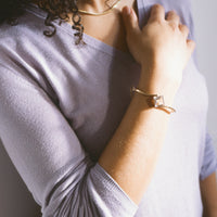 A close-up lifestyle shot of a person wearing a lavender long-sleeve knit top, a gold chain necklace, and a gold bracelet with a square bezel-set stone.