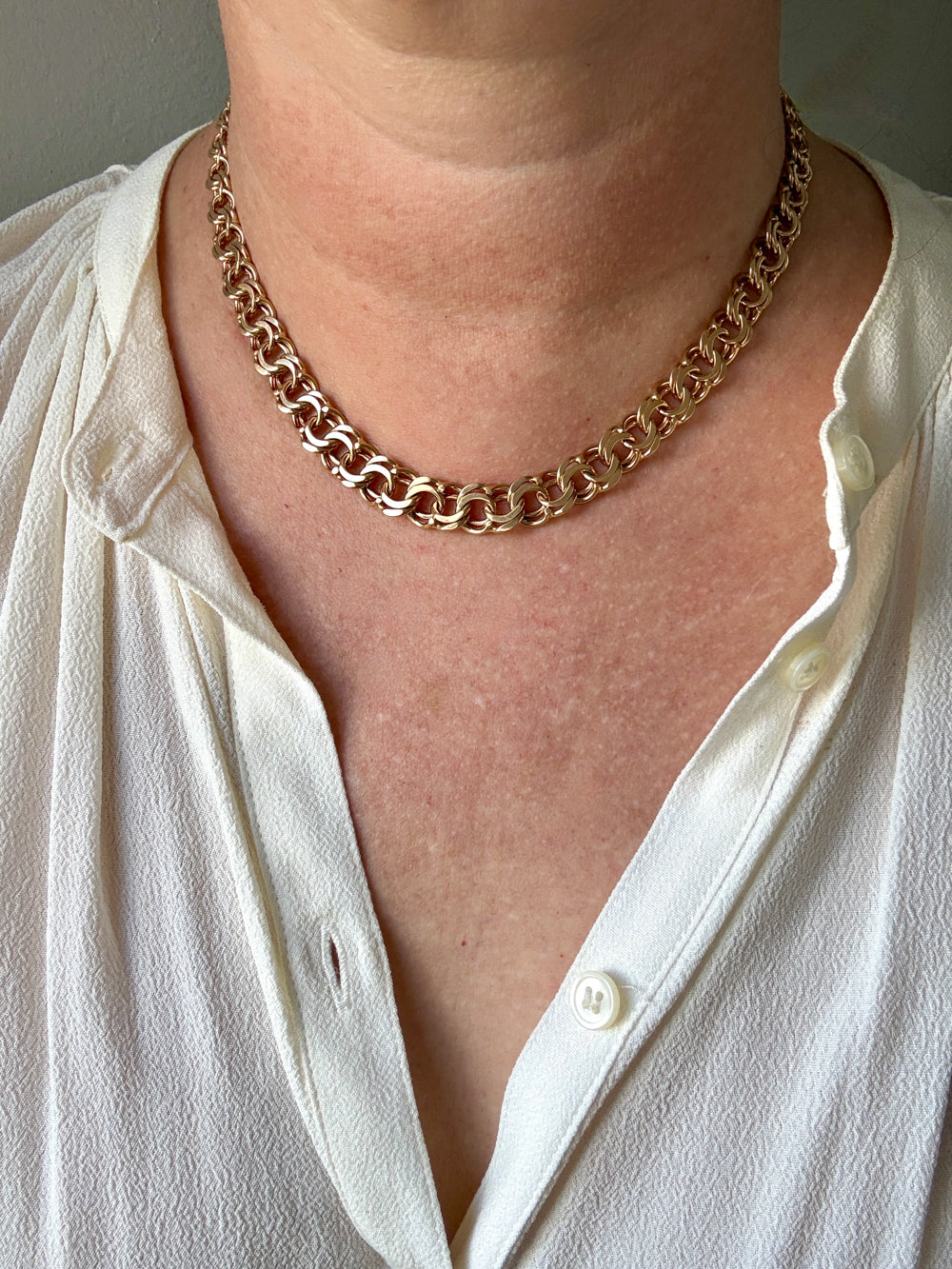 A close-up of a person wearing a chunky 18k gold chain necklace over a white button-up shirt.