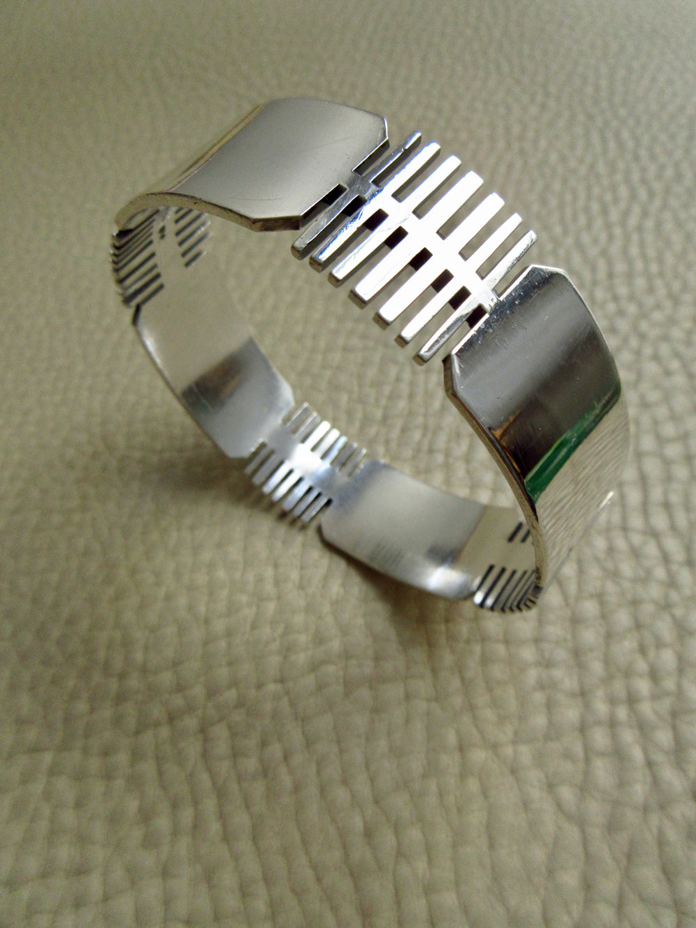 A polished silver bangle bracelet with geometric, ribbed cutout patterns, displayed on a textured beige leather surface.