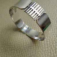 A polished silver bangle bracelet with geometric, ribbed cutout patterns, displayed on a textured beige leather surface.