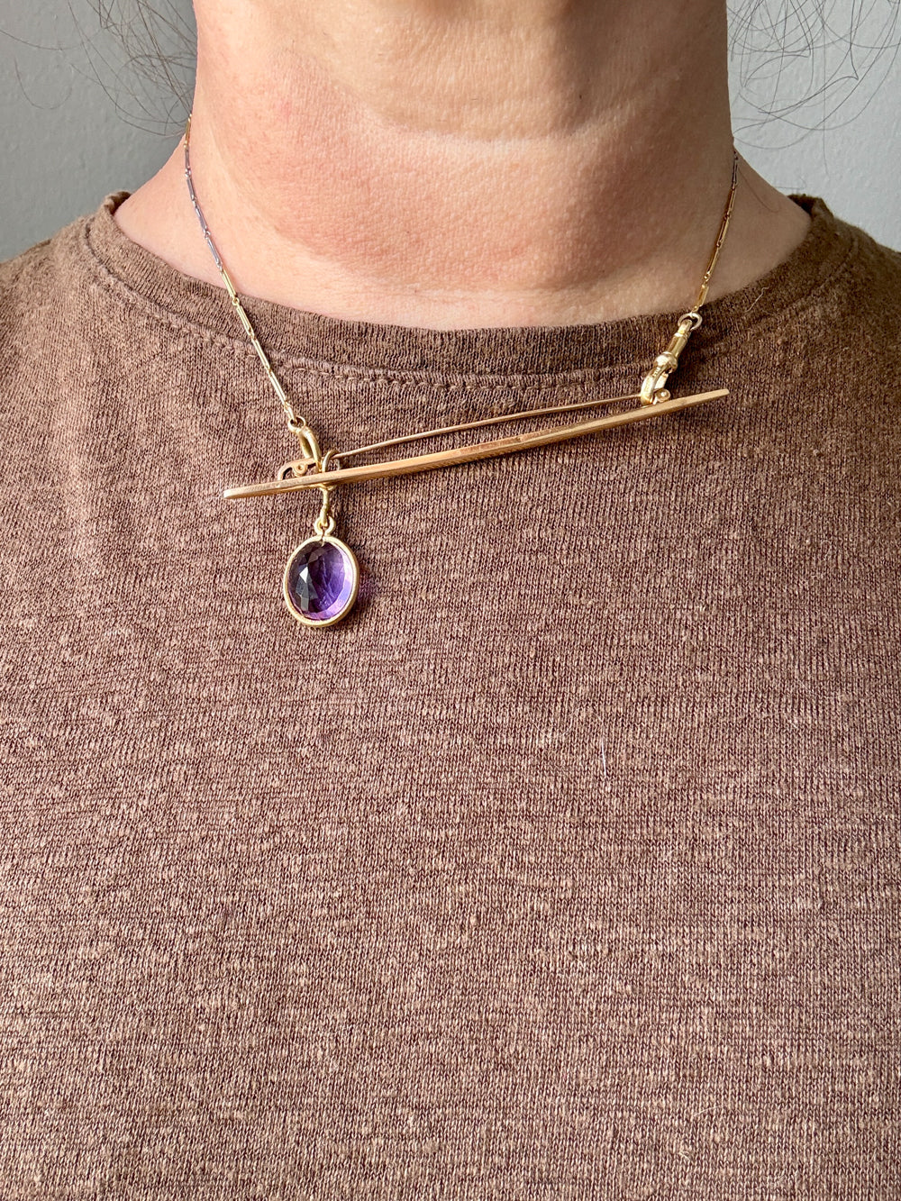 Gold necklace with a purple gemstone pendant worn by a person on a plain background