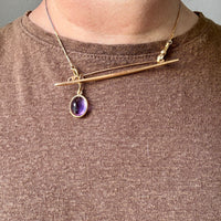 Gold necklace with a purple gemstone pendant worn by a person on a plain background