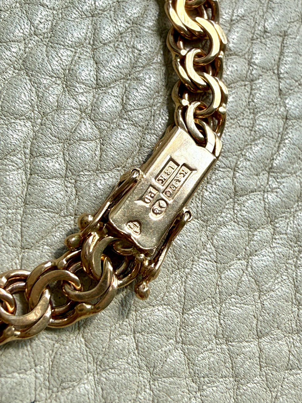 Close-up of a gold chain rectangular clasp with metal hallmarks, resting on a textured light gray surface.