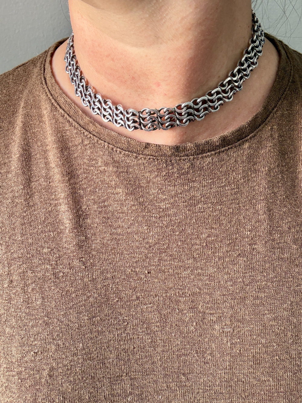 A close-up of a person's neck and upper chest wearing a chunky vintage silver chain choker with interlocking chain links, paired with a brown knit t-shirt.