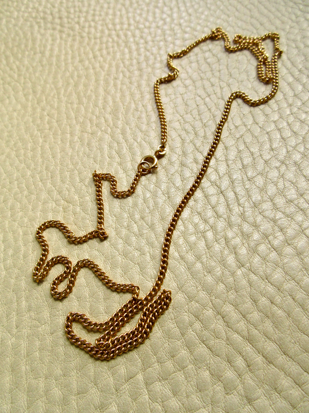 An 18k gold chain necklace with a curb link style, laid out on a textured beige leather surface.