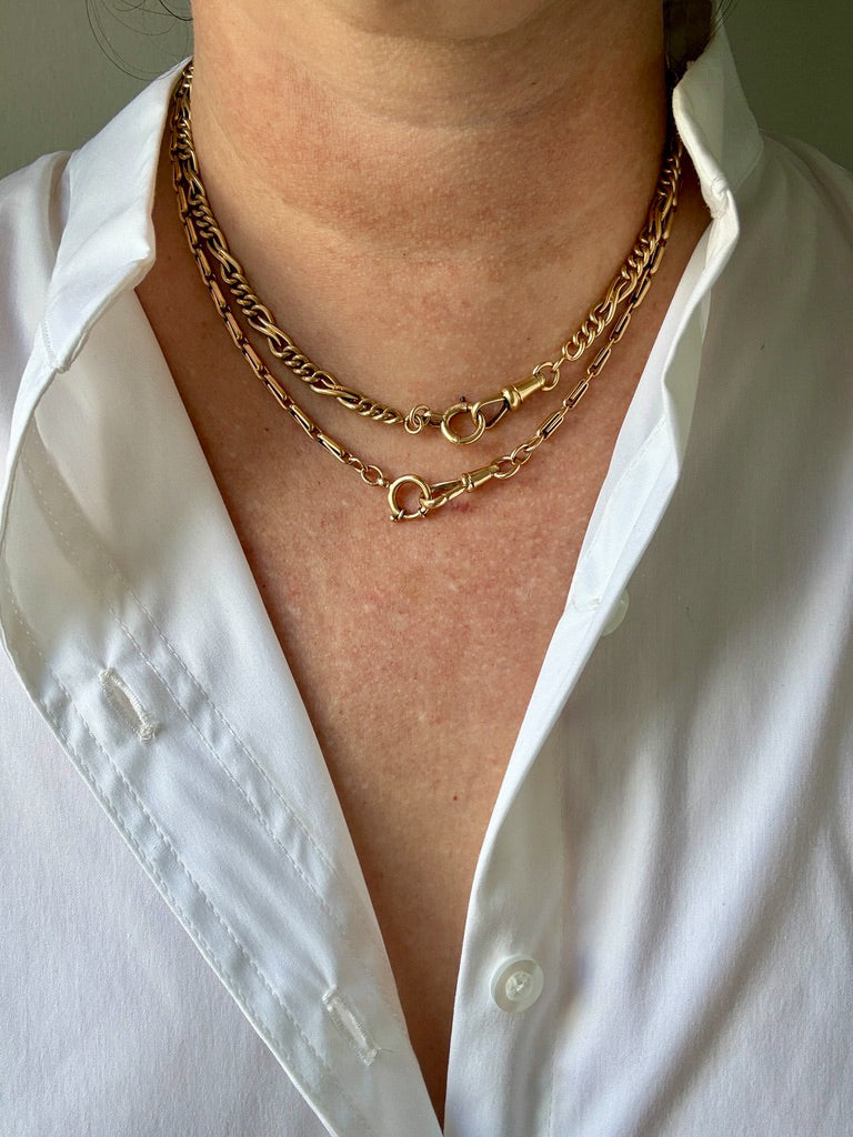 A close-up of a person wearing two layered 18k gold watch chain necklaces over a white button-down shirt.