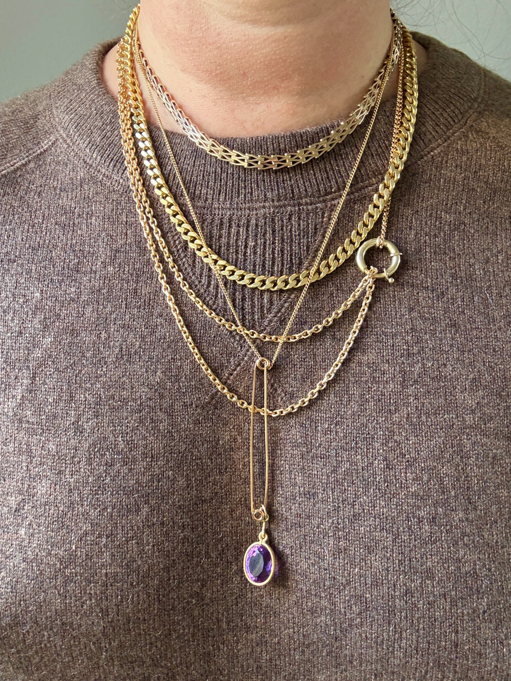 Person wearing multiple gold necklaces with a purple pendant on a brown sweater.