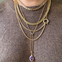 Person wearing multiple gold necklaces with a purple pendant on a brown sweater.