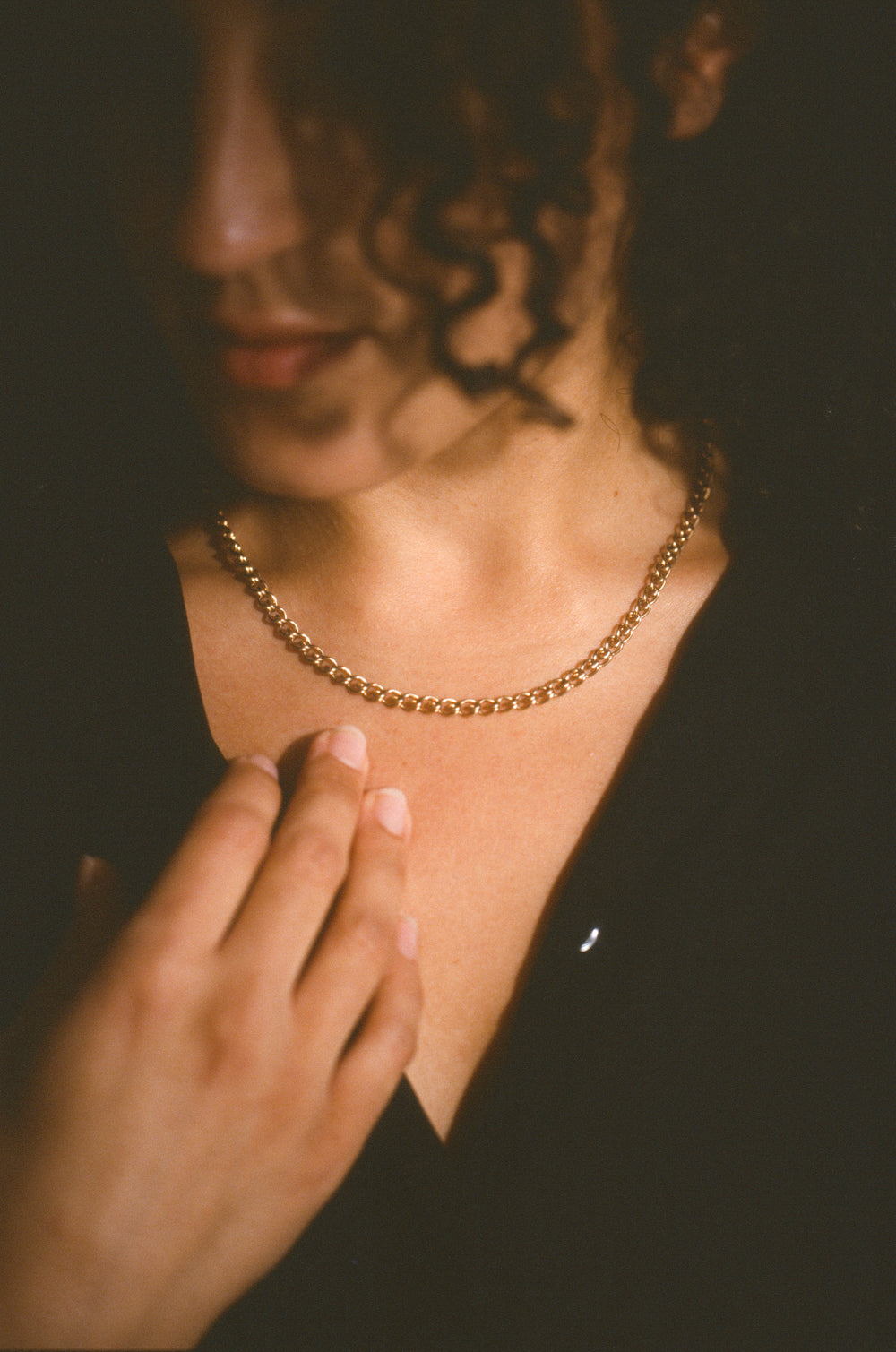 A close-up lifestyle shot of a person wearing a delicate gold chain necklace, with their hand resting near their collarbone against a dark background.