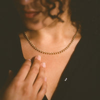 A close-up lifestyle shot of a person wearing a delicate gold chain necklace, with their hand resting near their collarbone against a dark background.