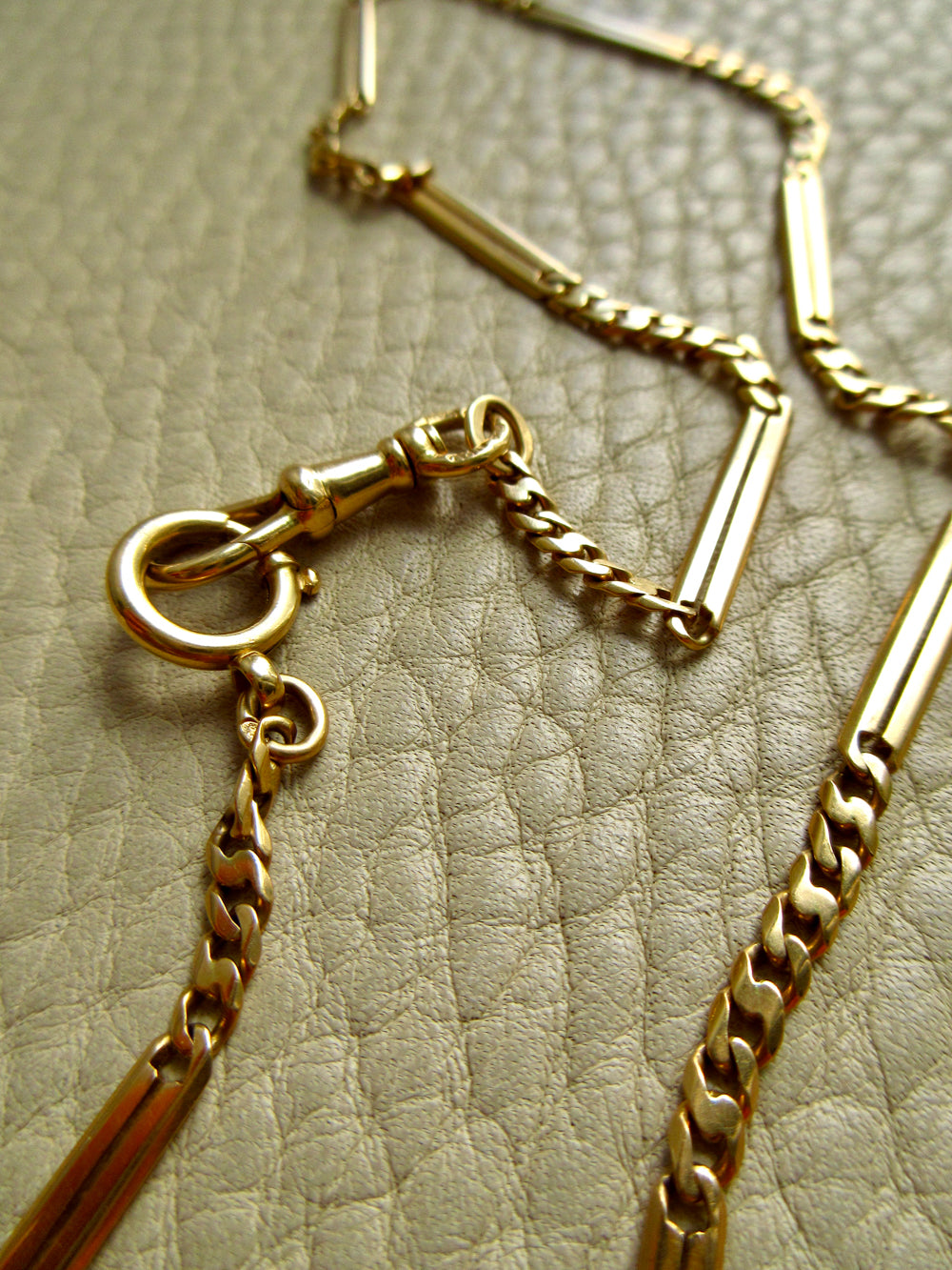 A close-up of an antique 18k gold chain necklace with watch chain clasps, laid on a textured light beige surface.