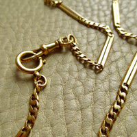 A close-up of an antique 18k gold chain necklace with watch chain clasps, laid on a textured light beige surface.