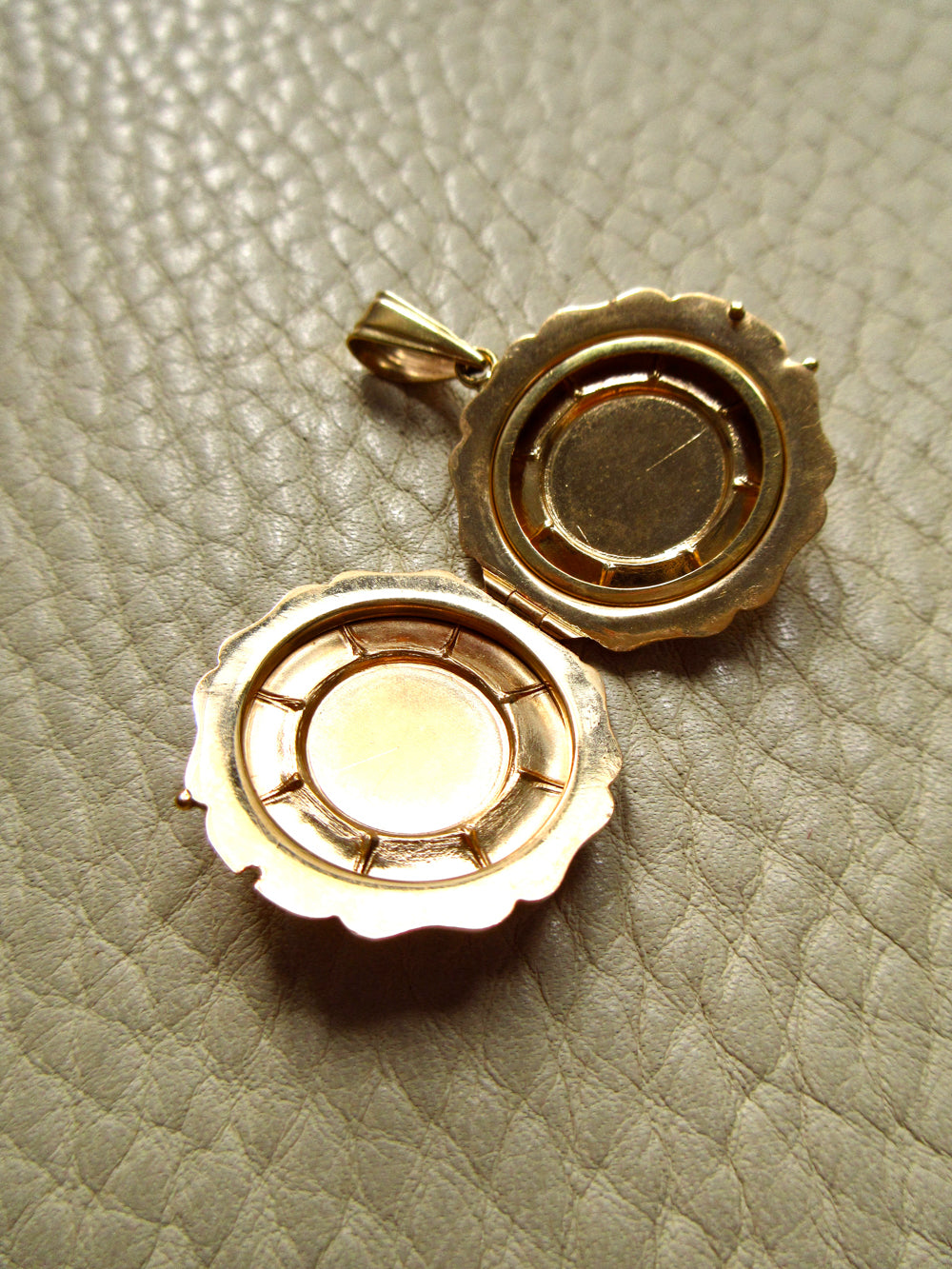 An open gold locket pendant with a scalloped edge, displayed on a textured light beige leather surface.