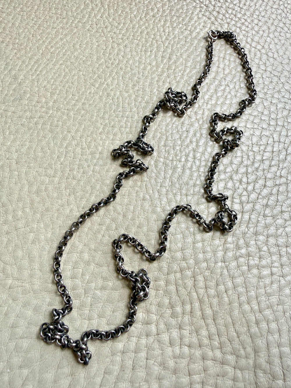 A patina'd silver chain necklace with a textured, oxidized finish, laid out on a light beige pebbled leather background.
