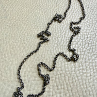 A patina'd silver chain necklace with a textured, oxidized finish, laid out on a light beige pebbled leather background.