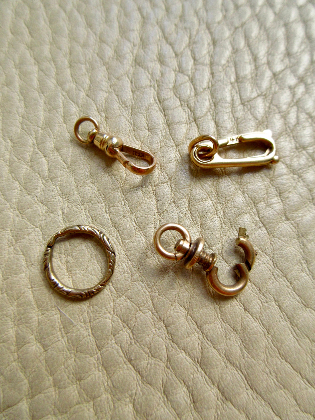 Four small gold jewelry findings arranged on a textured beige leather surface