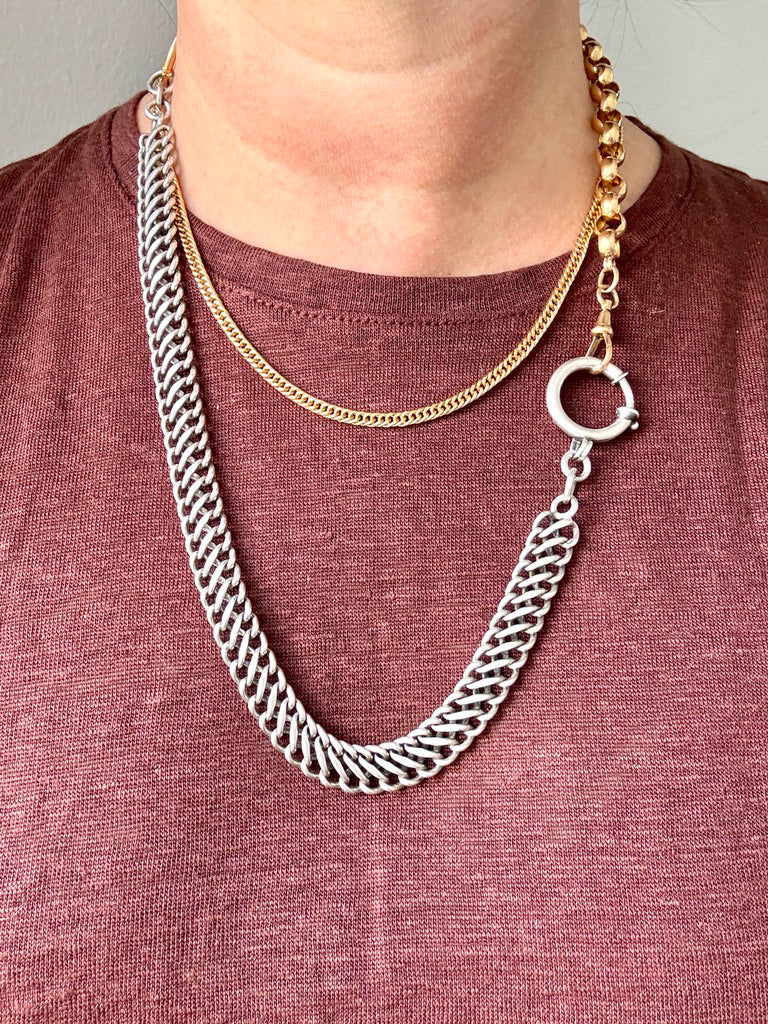 A close-up of a person wearing a layered necklace set: a thick silver curb chain draped over a gold chain, paired with a burgundy heathered t-shirt.