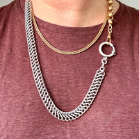 A close-up of a person wearing a layered necklace set: a thick silver curb chain draped over a gold chain, paired with a burgundy heathered t-shirt.
