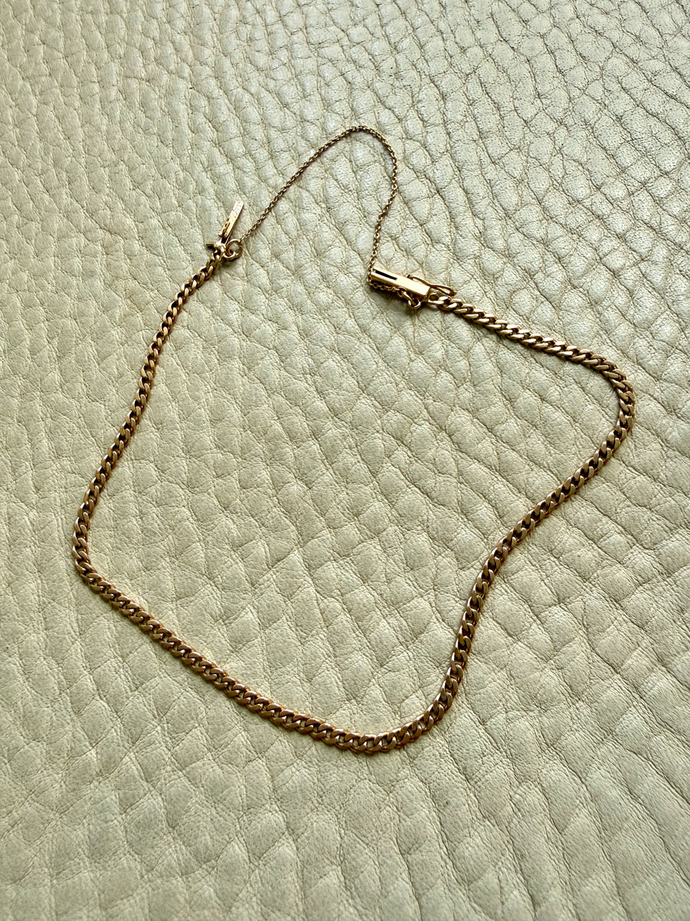 An 18k gold chain bracelet with a box clasp and safety chain, laid out on a textured off-white leather surface.