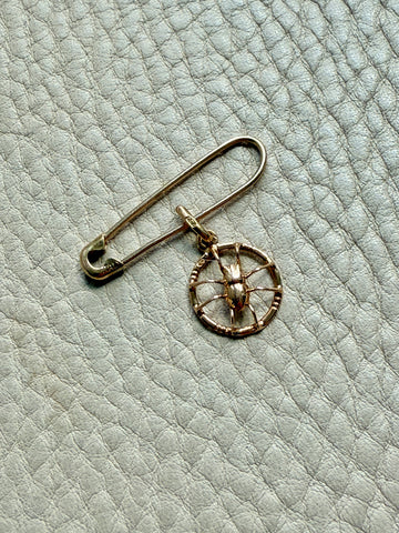 Gold safety pin with a circular spider pendant on a light gray textured background