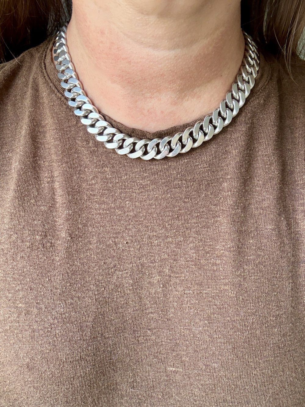 A close-up of a person wearing a chunky silver Cuban link chain necklace over a brown knit top.