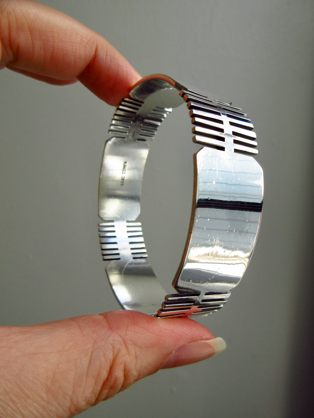 A hand holding a polished silver bangle bracelet with horizontal slit details on the inner band, shown against a plain gray background.