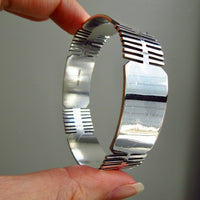 A hand holding a polished silver bangle bracelet with horizontal slit details on the inner band, shown against a plain gray background.