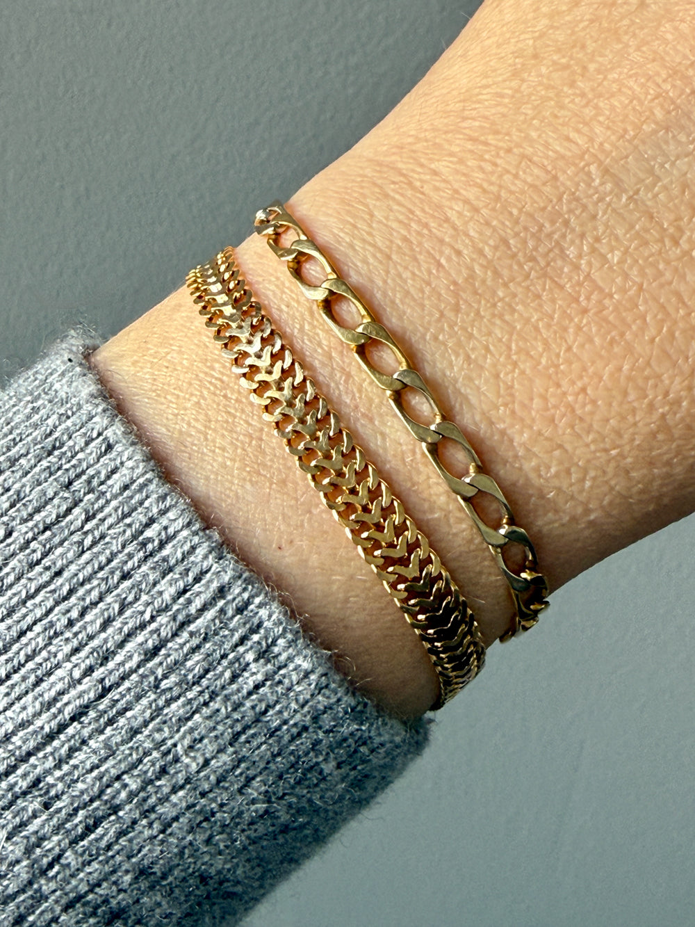 Two 18k gold vintage chain bracelets stacked on a person's wrist, with a gray knit sweater cuff visible at the lower left against a muted gray background.