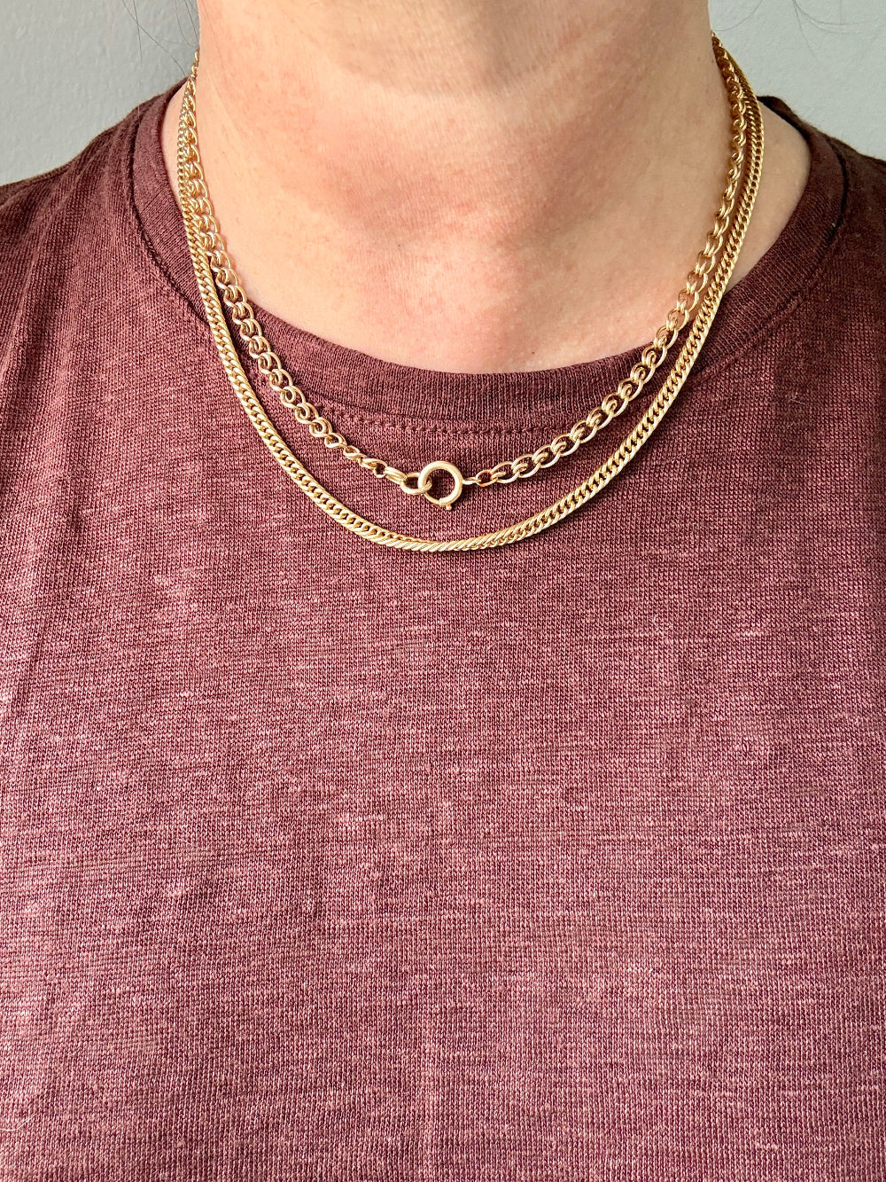 A close-up of a person's neck wearing two layered gold chain necklaces over a heathered mauve-brown t-shirt.