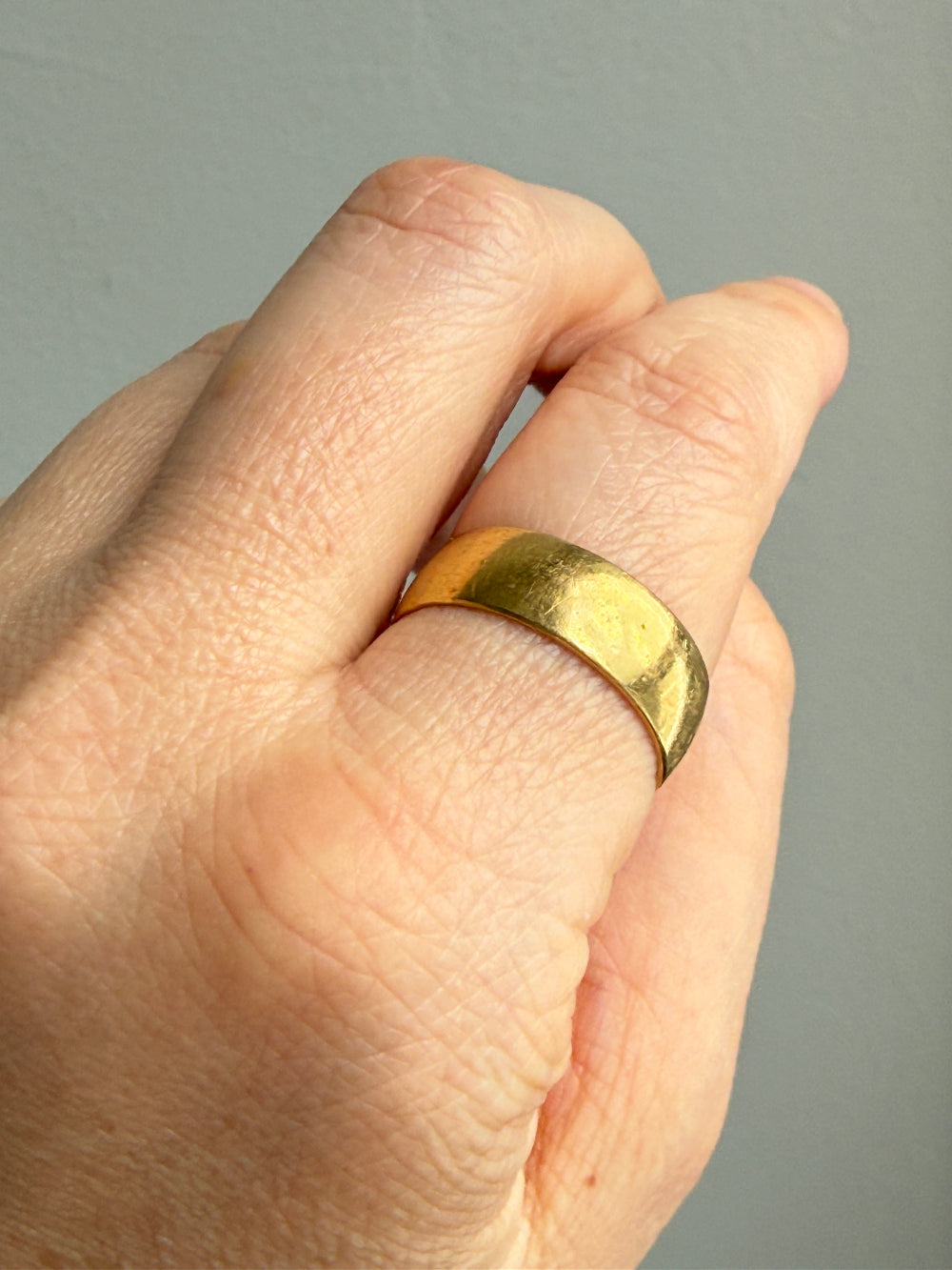 A close-up of a hand wearing a simple, wide gold band ring against a plain gray background.
