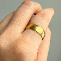 A close-up of a hand wearing a simple, wide gold band ring against a plain gray background.