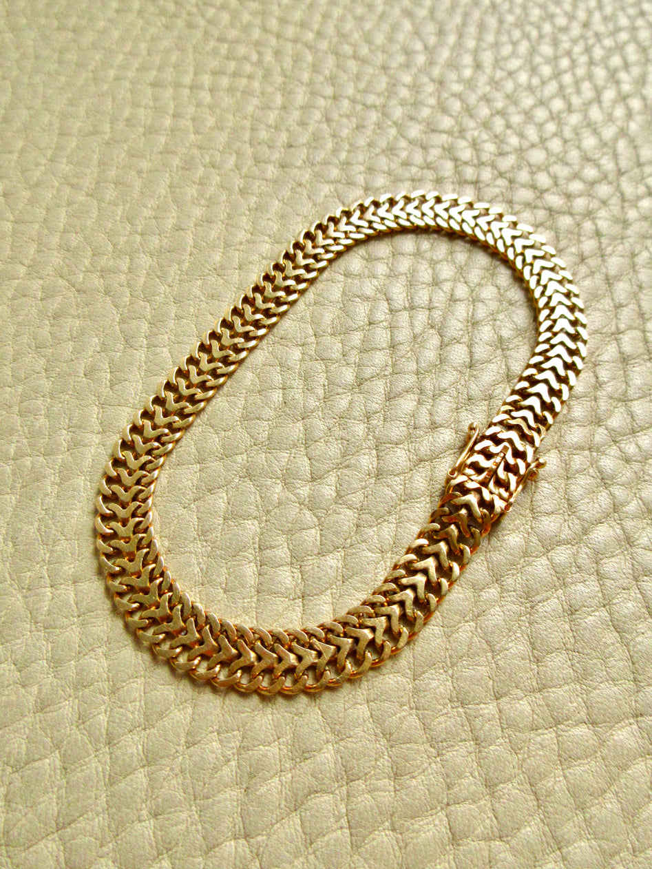 A vintage 18k gold herringbone chain braceley with a woven, braided link design, arranged in an oval shape on a textured beige leather background.