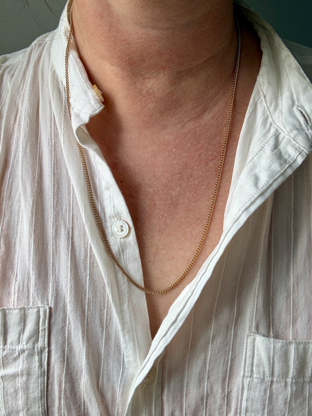 A close-up of a person's neck and chest wearing a delicate gold chain necklace over an open white linen button-up shirt.