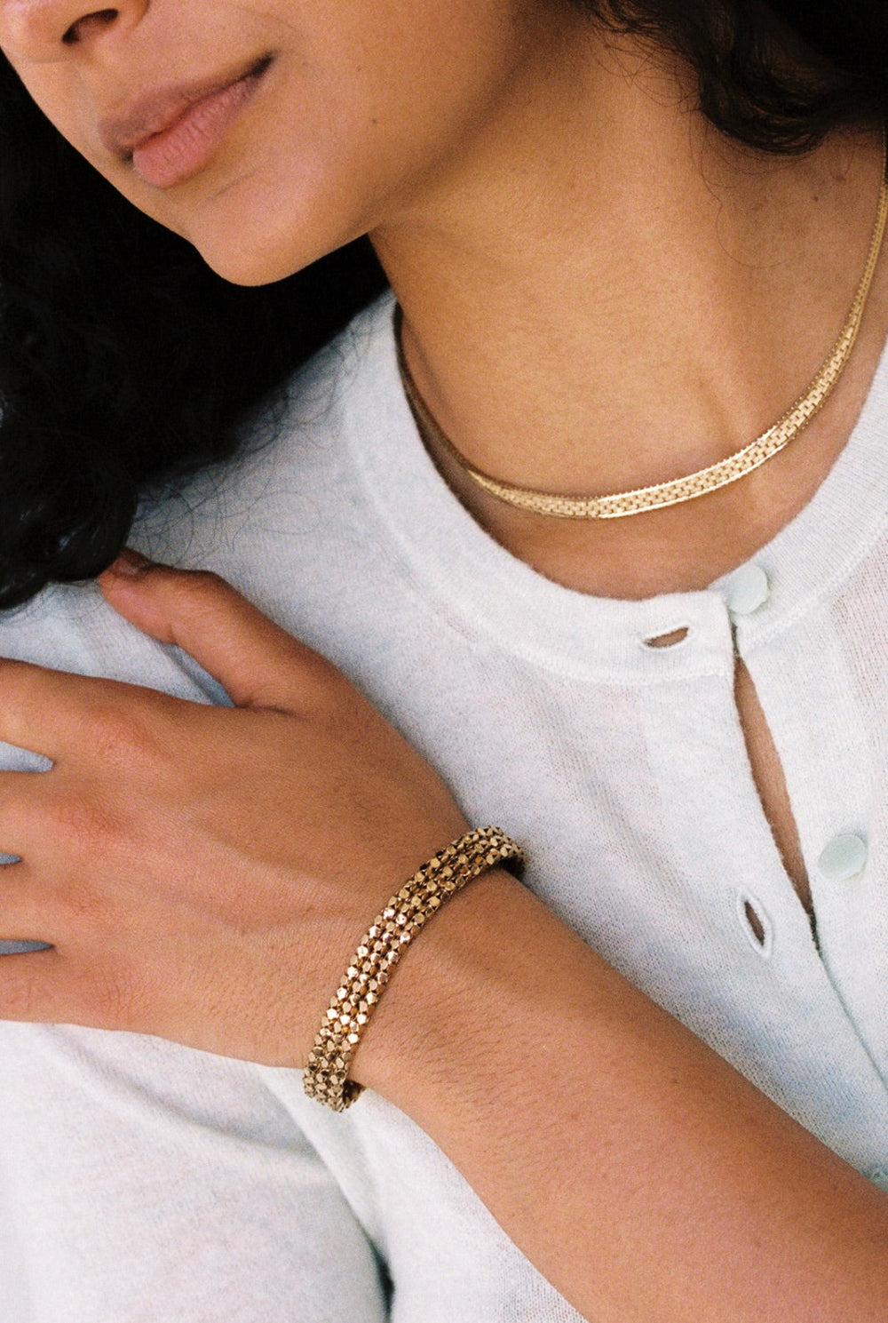 A close-up lifestyle shot of a model wearing a gold flat link necklace and an 18k gold honeycomb link bracelet on her wrist, paired with a white button-up shirt. penelopepenelope.com