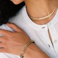 A close-up lifestyle shot of a model wearing a gold flat link necklace and an 18k gold honeycomb link bracelet on her wrist, paired with a white button-up shirt. penelopepenelope.com