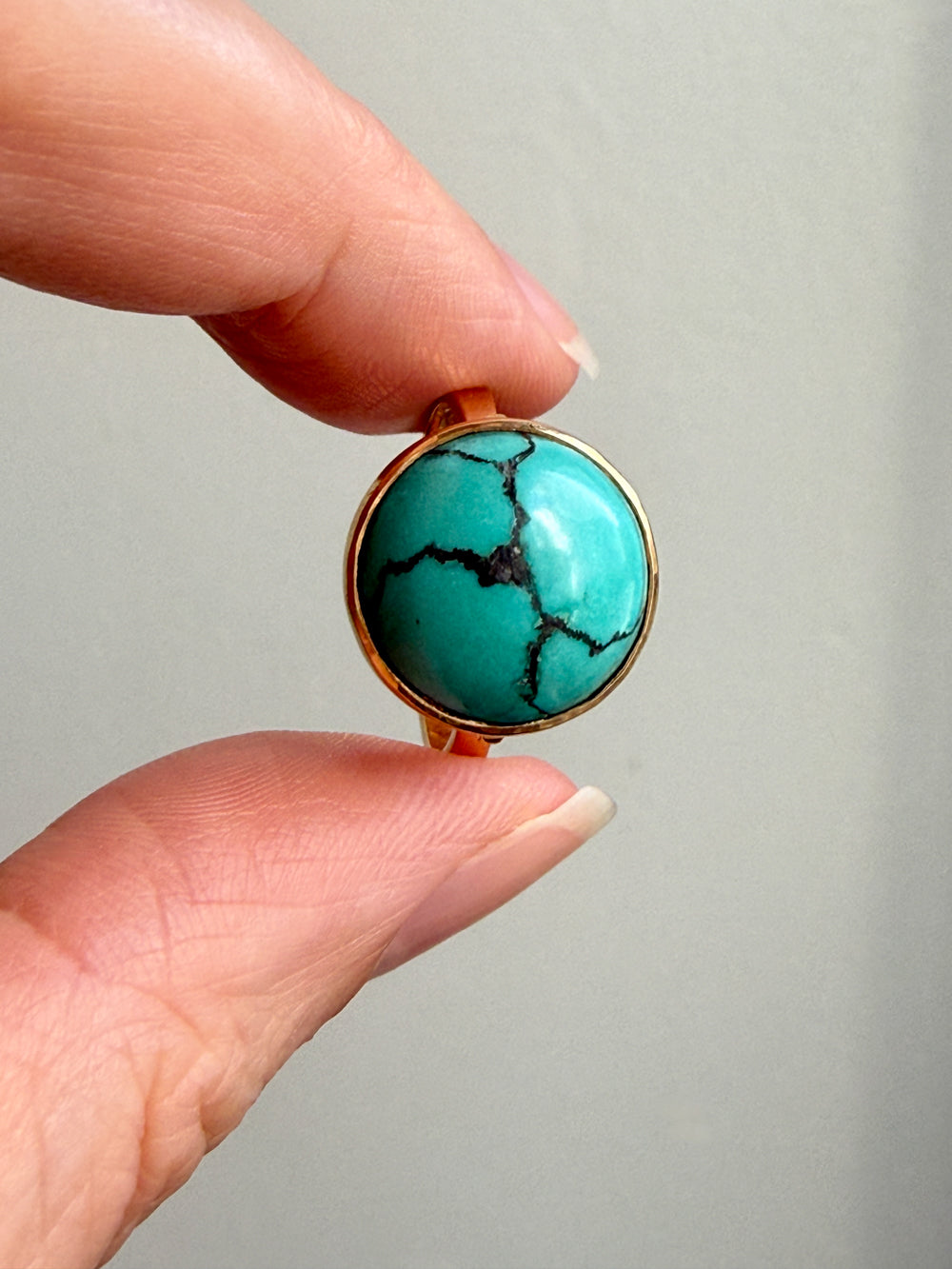 A close-up of a hand holding a round turquoise stone ring with a gold bezel setting against a plain light gray background.
