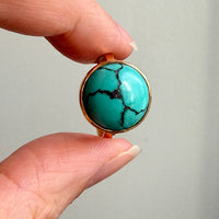 A close-up of a hand holding a round turquoise stone ring with a gold bezel setting against a plain light gray background.
