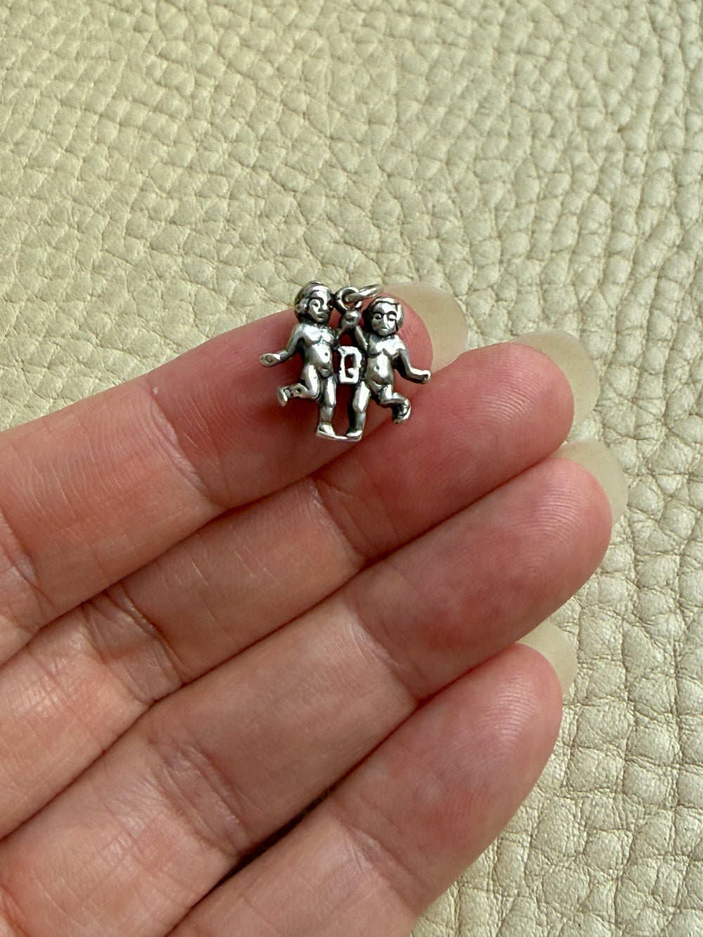 A small silver charm depicting two gemini figures, held between a person's fingers against a textured beige background.